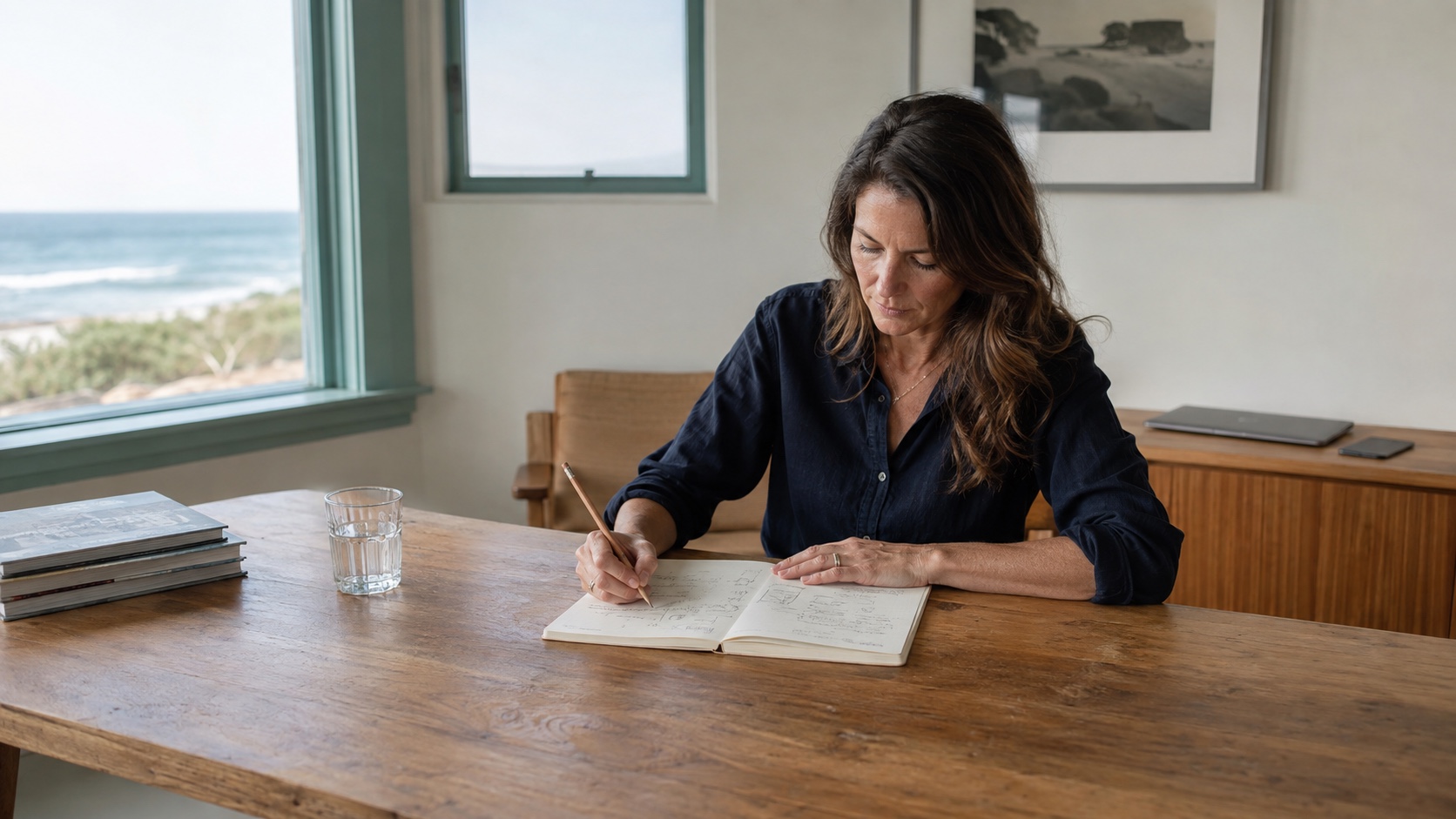 A woman concentrating on a paper project plan at a coastal studio table while her closed laptop and phone remain out of reach